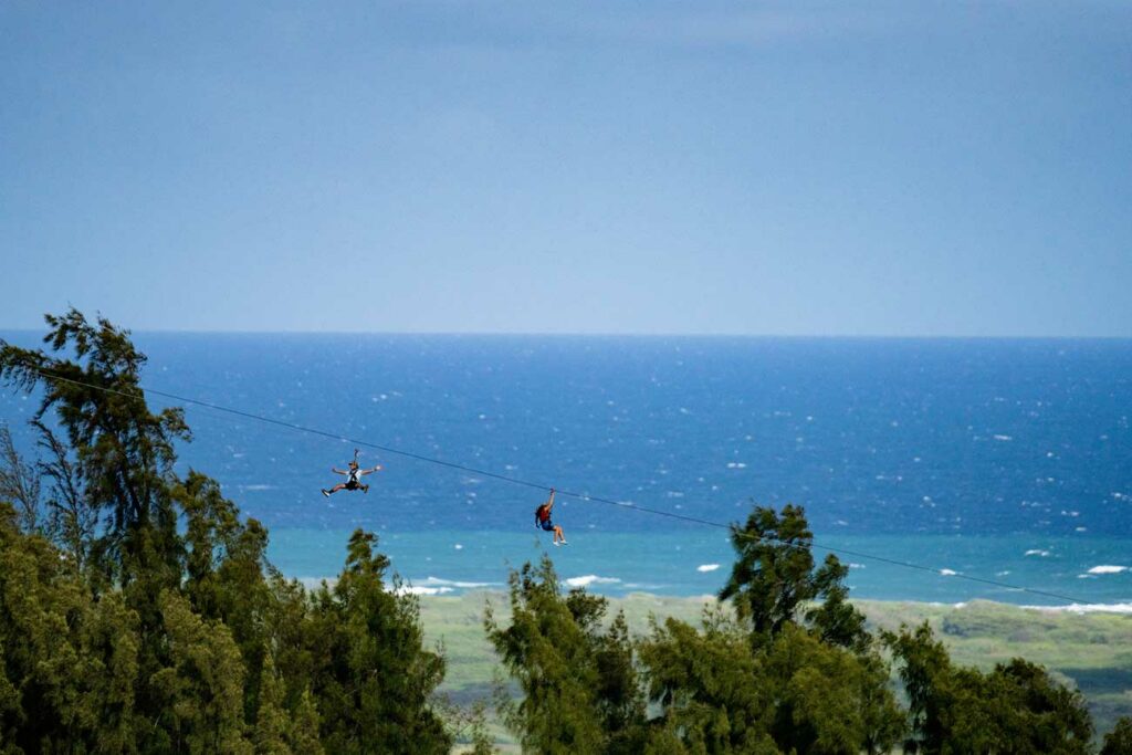 Two people ziplining on dual ziplines across a view of Oahu's North Shore