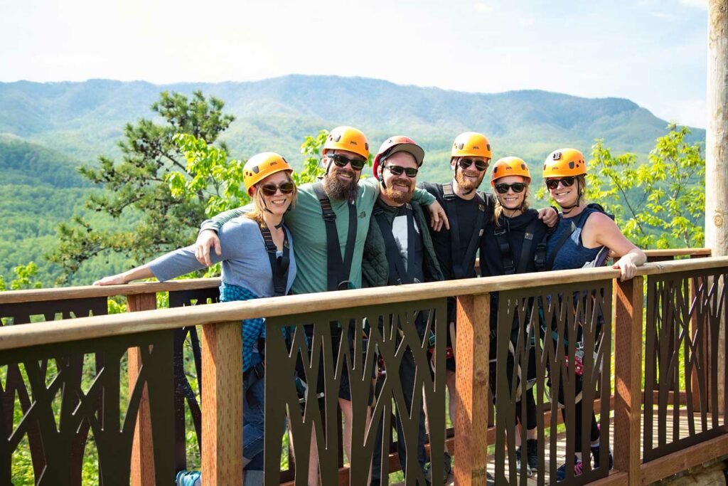 A smiling group poses in their harnesses and helmets on a platform against the backdrop of the Smoky Mountains