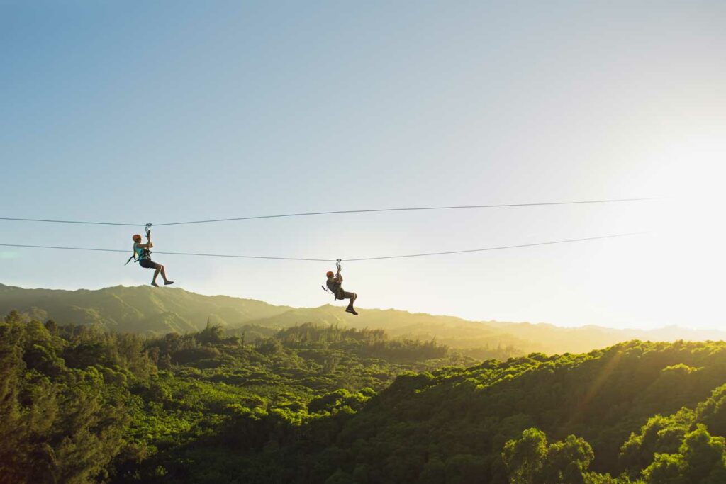 zipliners cross the sky above lush green mountaintops at golden hour in Hawaii